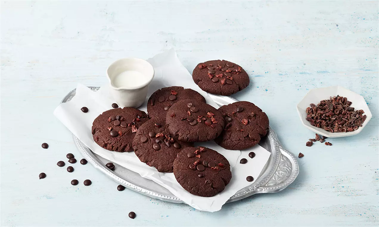 Кръгли веган бисквитки тъмнокафяви и плътни с поръски шоколадови капки върху бял лист хартия в изпъкнала метална чиния