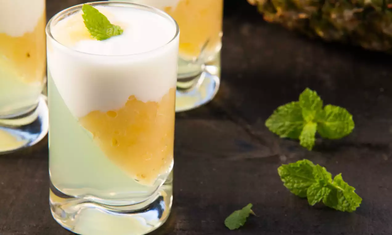 Mini taça de vidro transparente com creme branco encorpado sobre purê amarelo com textura granulada, decorada com folha de hortelã