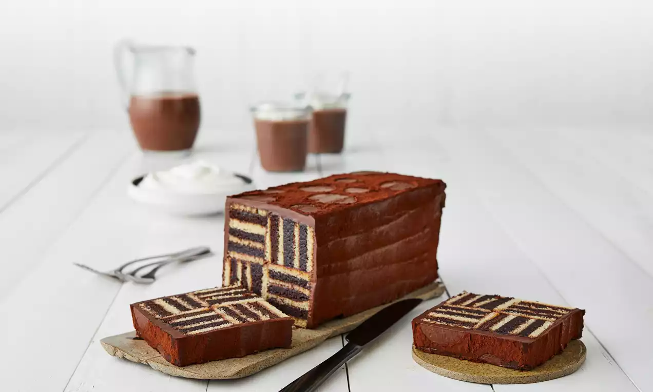 Rektangulær sjokoladetrekkkake med mønster av blå, gule, røde og hvite lag, servert på fat med kniv og tilbehør i bakgrunnen