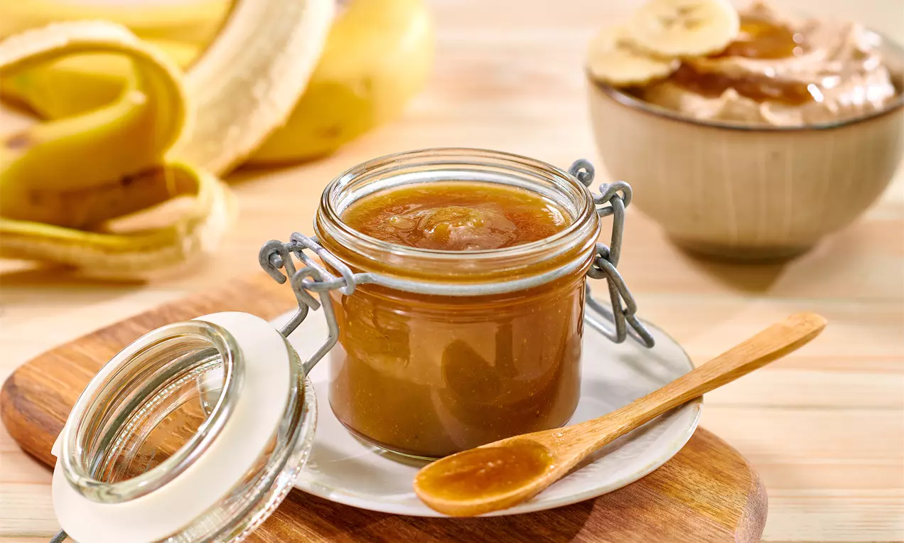 Златисто-кафяв карамелен крем с гладка и плътна текстура в прозрачен буркан с отворен капак, дървена лъжичка и фон с банани
