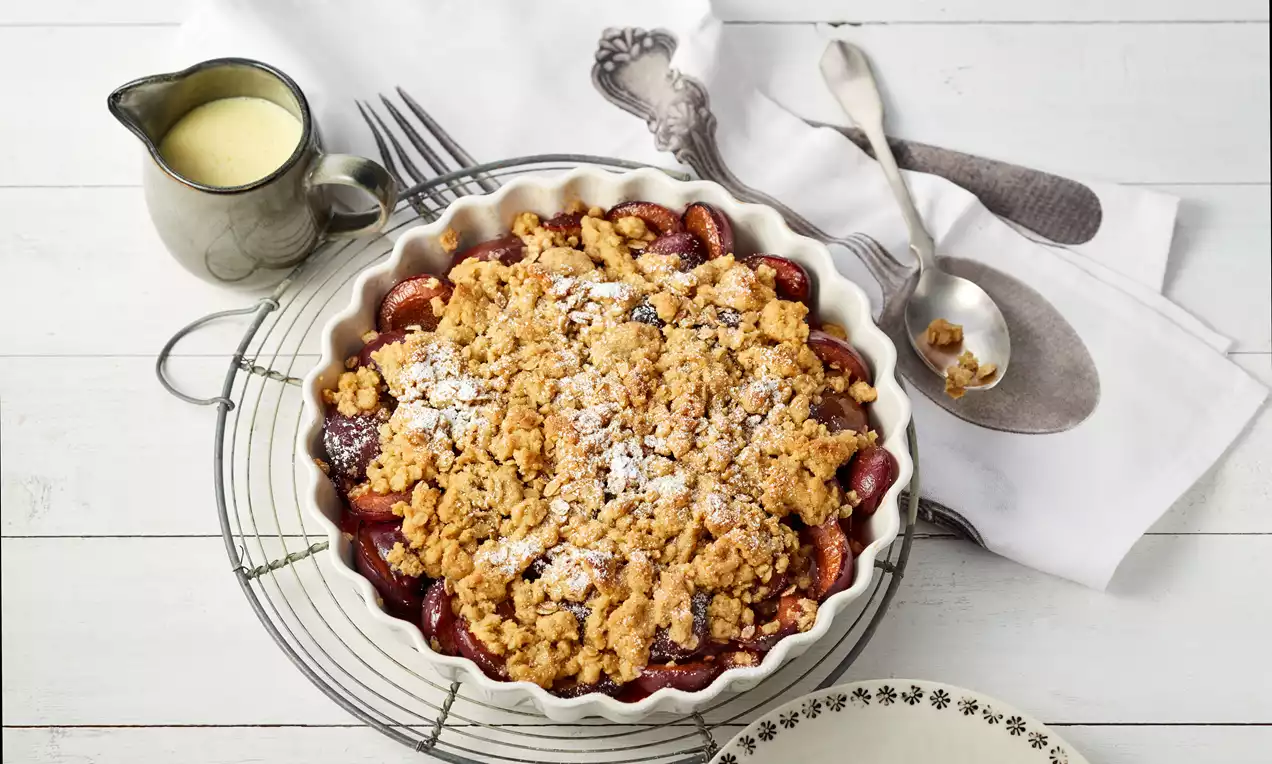 Zwetschgen-Crumble in einer weißen Auflaufform mit goldbraunen Streuseln und saftigen dunkelvioletten Zwetschgen, serviert mit Vanilleeis