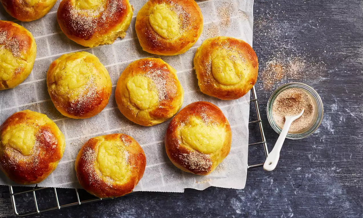 Saffransbullar med gyllene yta och ljus vaniljkräm i mitten, pudrade med kanelsocker, ligger på bakplåtspapper med burk och sked bredvid