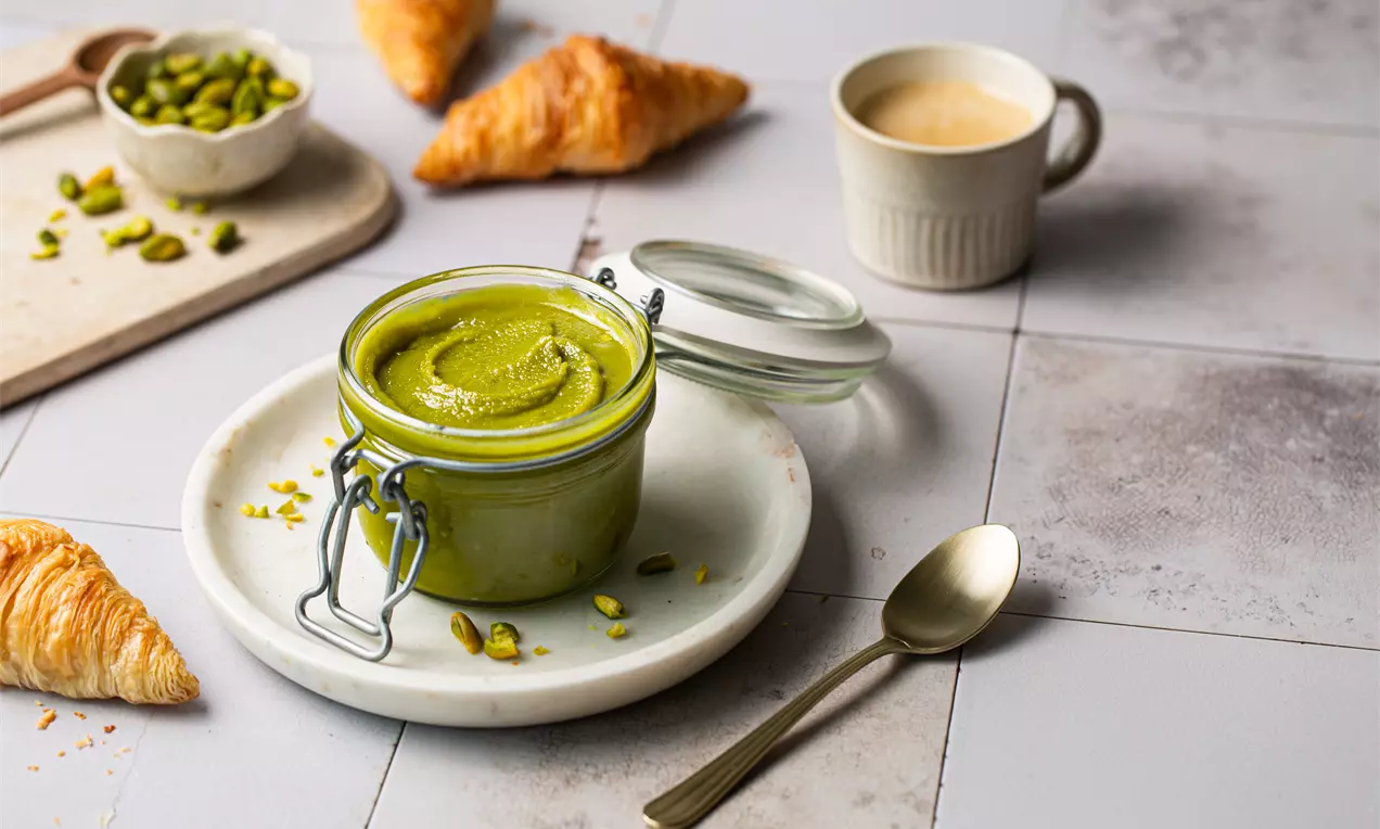 Lysegrønn pistasjkrem i glass med metallklemme på hvit tallerken med skje, pistasjnøtter, croissanter og kopp på flismønstret bord