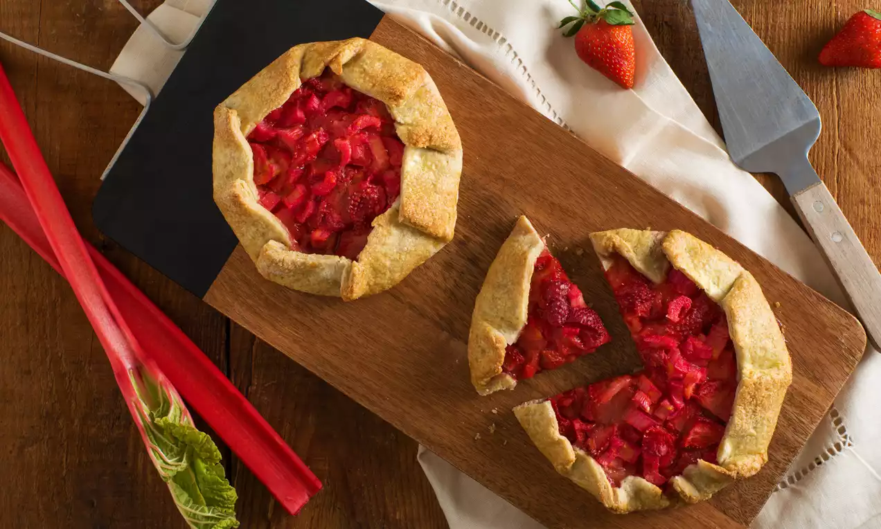 Golden-brown rustic galettes with crimped edges, filled with vibrant red strawberry and rhubarb chunks on a wooden cutting board