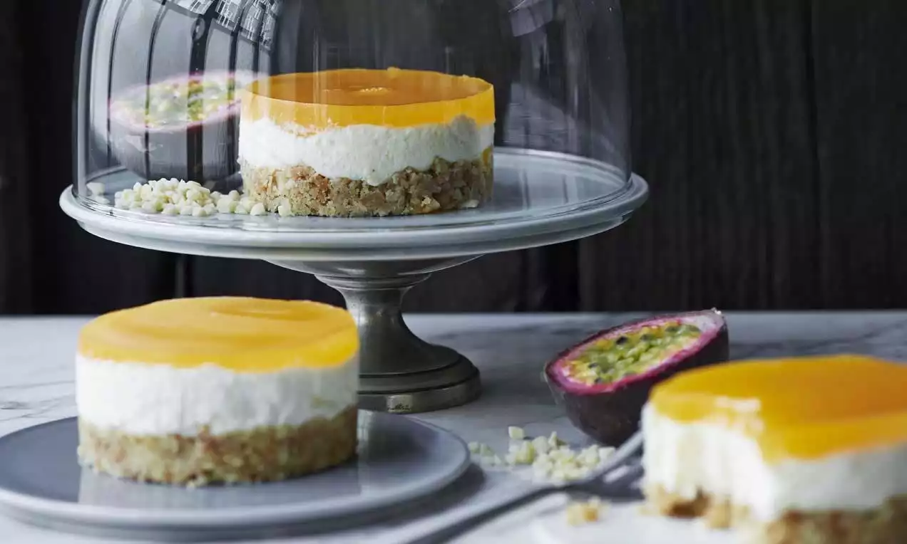 Runde porsjonsostekaker med gul pasjonsfruktgelé, hvitt kremet fyll og grov kjeksbunn servert på små asjetter og et kakefat i glass