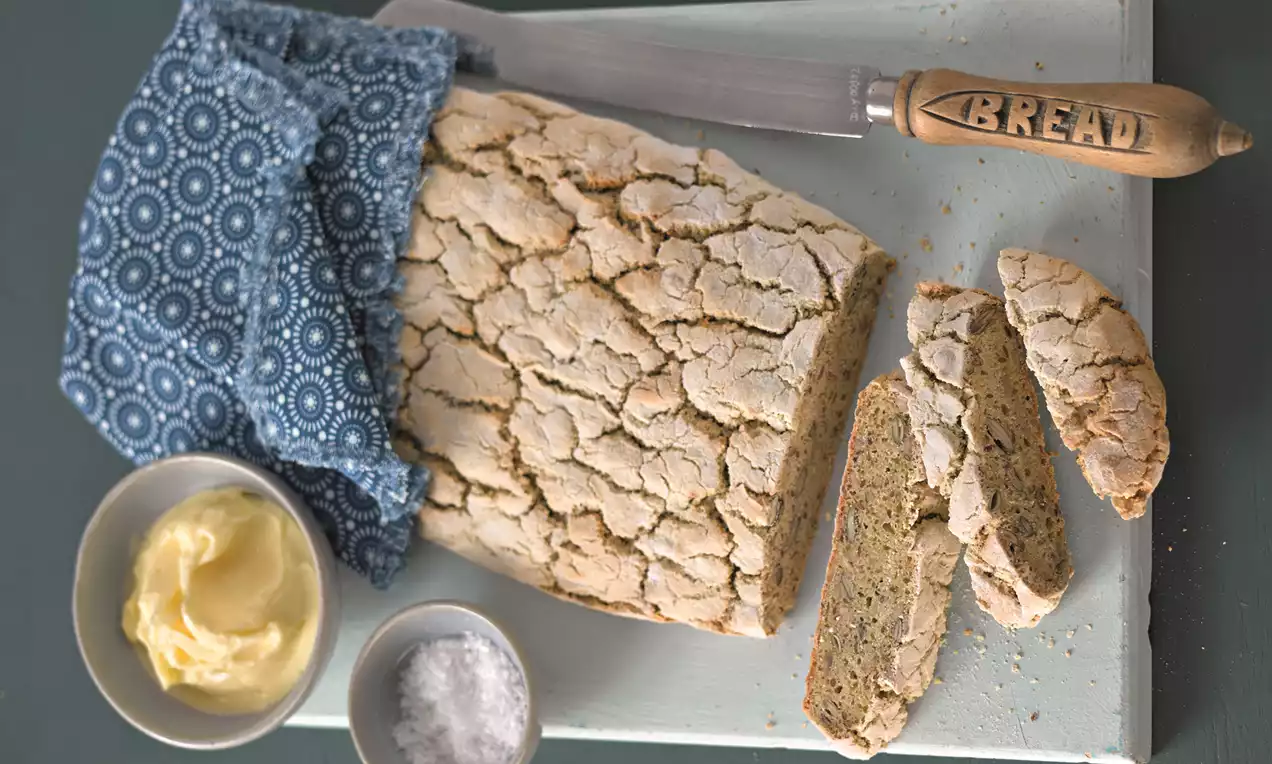 Glutenfreies Brot mit rissiger, goldbrauner Kruste, in Scheiben geschnitten auf einem hellen Brett mit Stofftuch, Messer, Butter und Salzschale