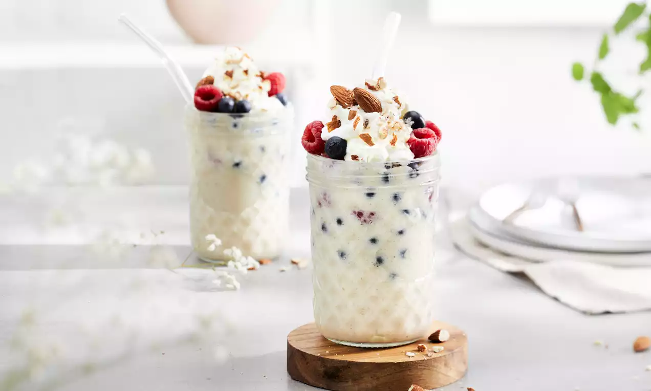 Batido cremoso de vainilla pálido con arándanos y frambuesas, terminado con nata, almendras troceadas y coco rallado en vasos de vidrio