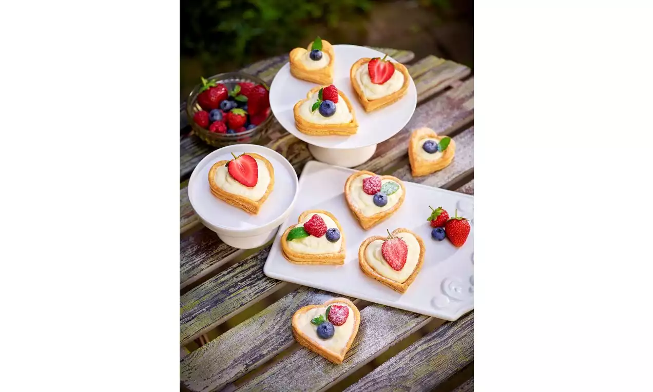 Feuilletés en forme de cœur dorés garnis de crème blanche et décorés de fraises, framboises, myrtilles et feuilles de menthe