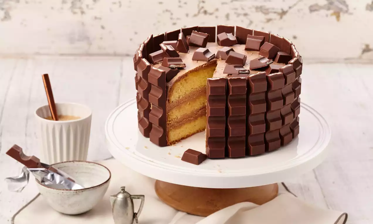 Runde Torte mit gelben Biskuitschichten, umhüllt von Kinder-Riegel®-Stücken, auf einer weißen Tortenplatte mit Karamellsoße und Deko