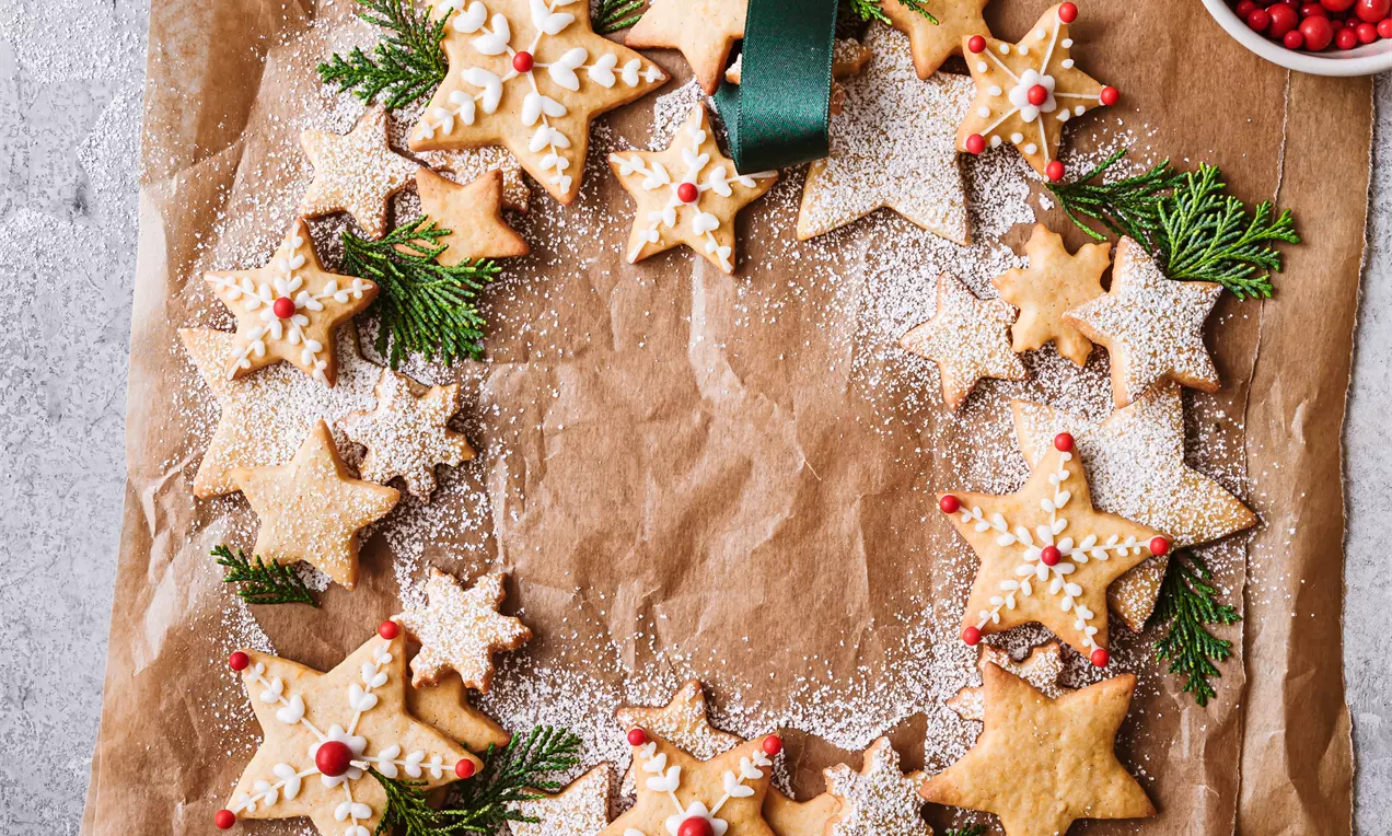 Picture - Star-shaped vanilla cookies with golden crisp edges, decorated with icing and berries, arranged in a wreath on brown parchment with fir sprigs
