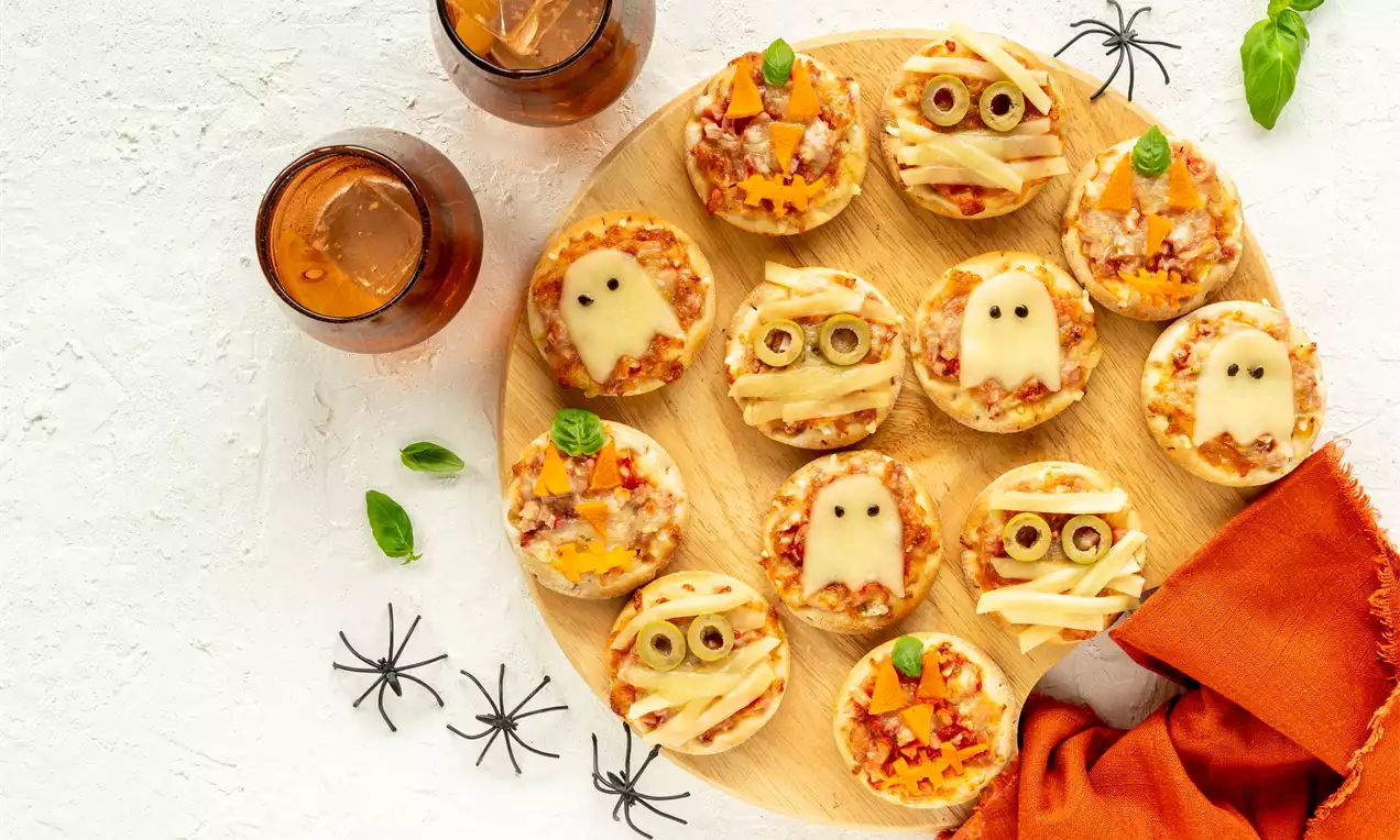 Petites pizzas rondes aux visages effrayants en fromages fondus et olives disposées sur une planche bois avec araignées en décor