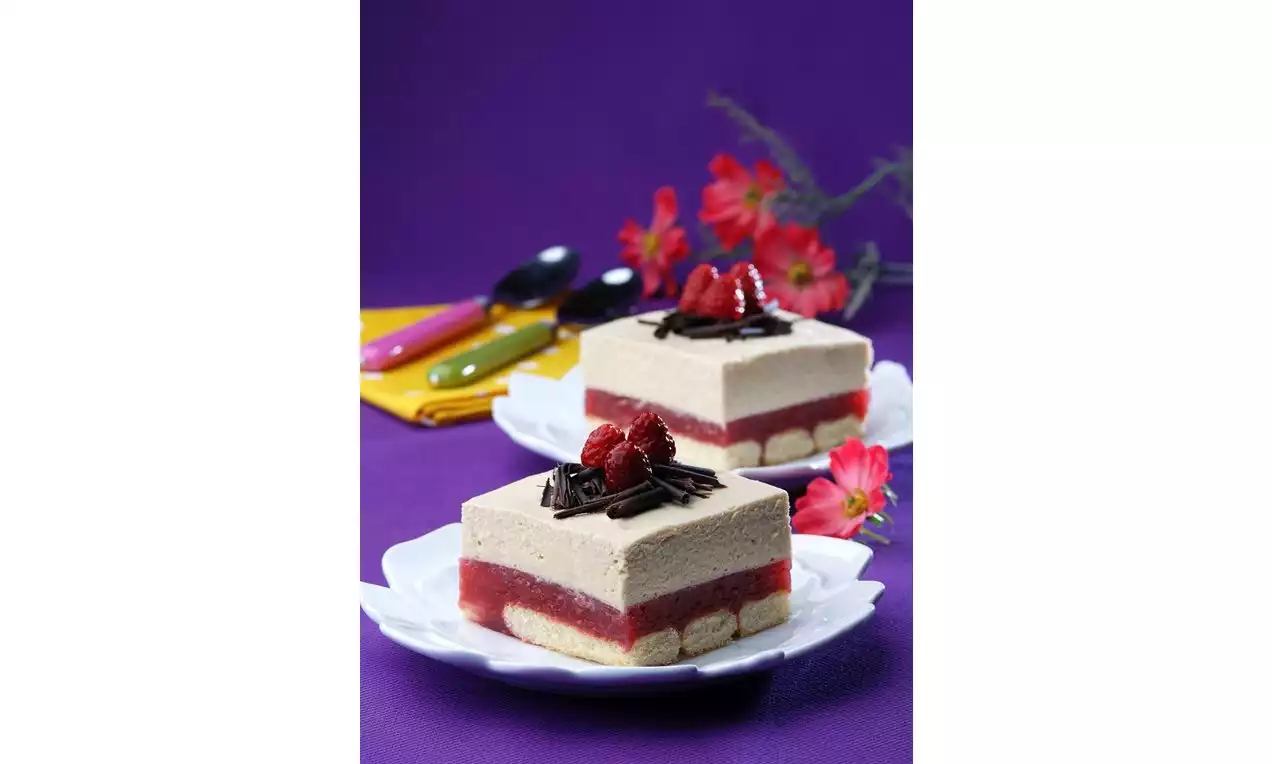 Alt tabanı katmanlı bisküvi, orta kısmı parlak kırmızı ahududu dolgu ve üst kreması açık kahverengi olan tiramisu, çikolata şeritleri ve taze ahududu ile süslenmiş, beyaz tabaklarda sunulmuş, mor bez zemin ve zarif kırmızı çiçek detaylarıyla dekorlanmış