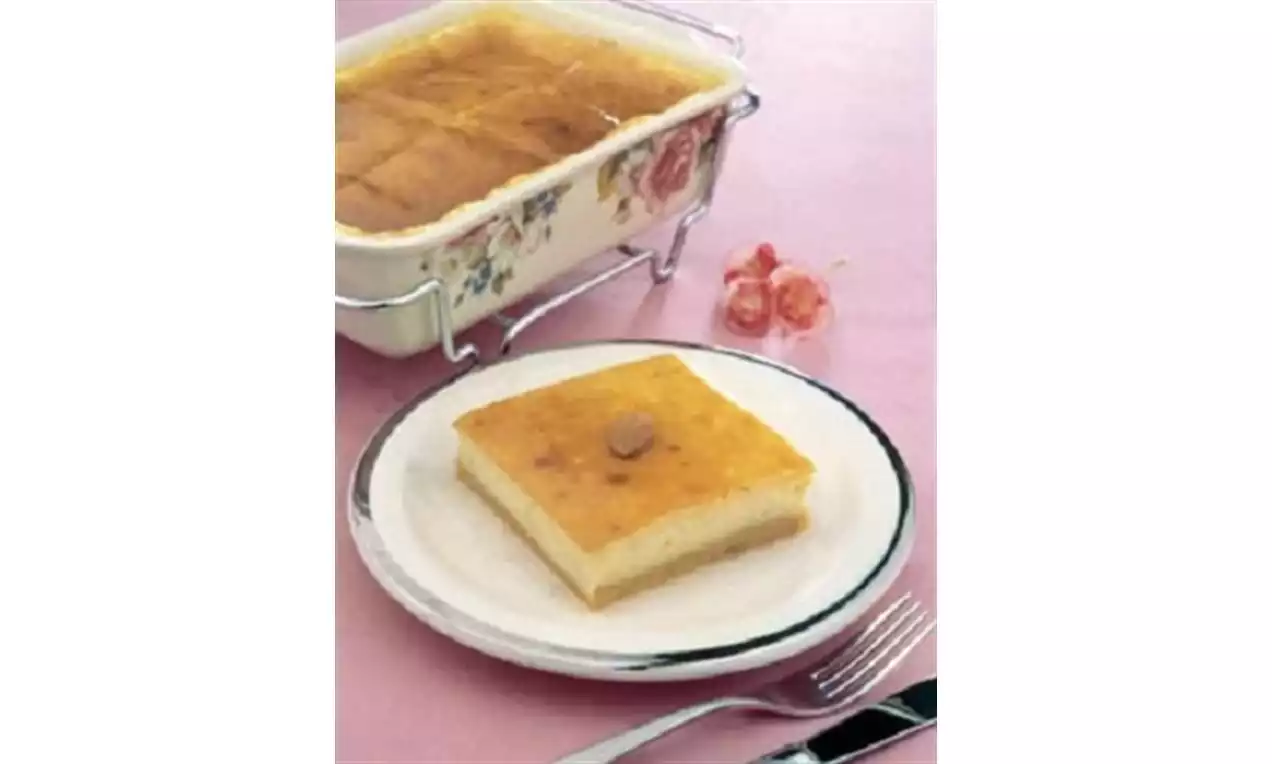 Kare dilimlenmiş, üzeri hafif kızarmış suruplu muhallebi beyaz tabakta sunulmuş, yanında çiçek desenli kabıyla tamamlanmış