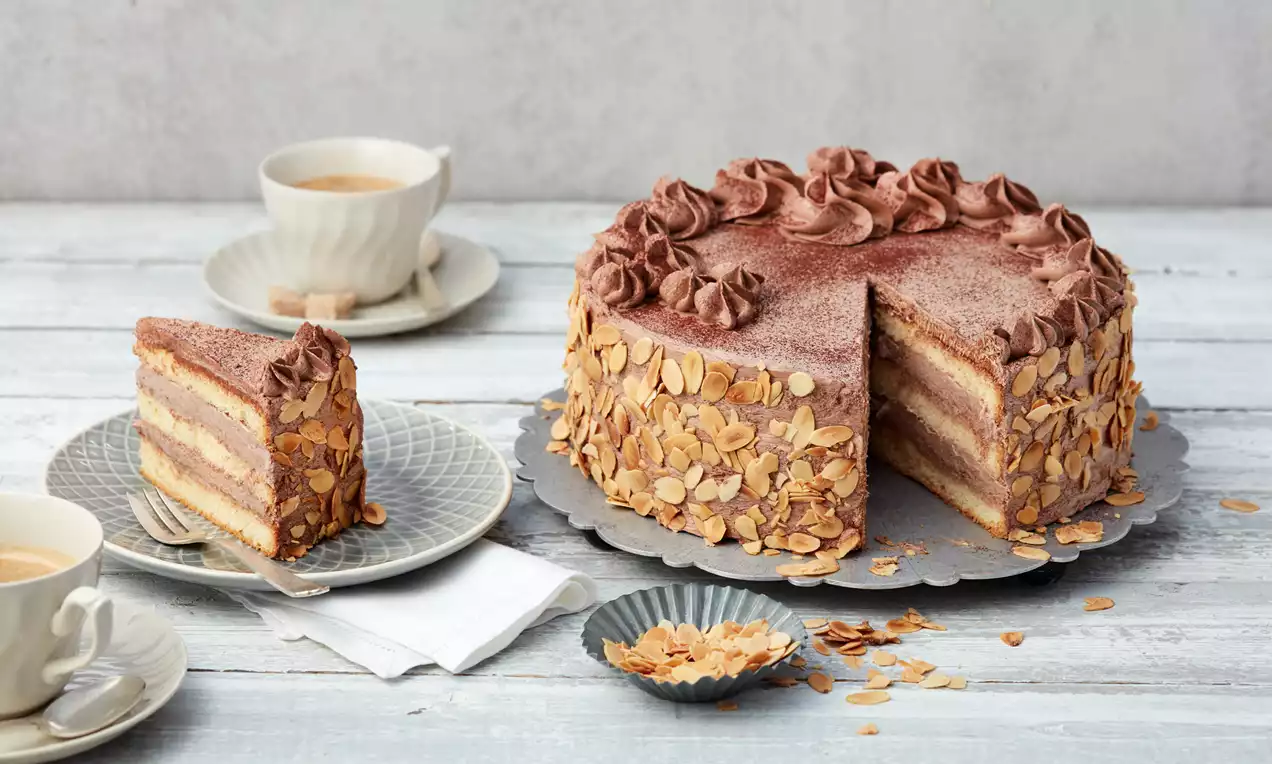 Buttercremetorte mit Schokoladencreme-Rosetten und mandelbesetztem Rand, daneben ein Stück auf grauem Teller und eine Tasse Kaffee